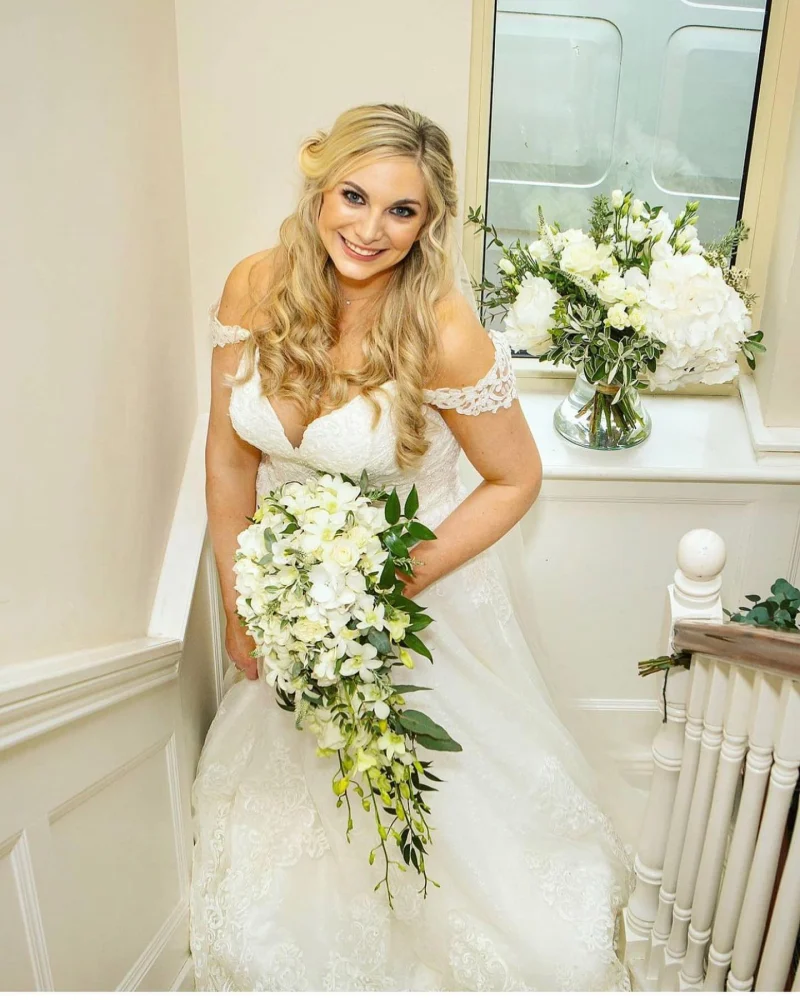 Bride standing at the bottom of the stairs, holding her bouquet - makeup by Nima